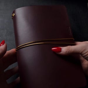 May include: A burgundy leather journal with a gold elastic closure. The journal is held by a person with red nail polish. The journal has a simple, elegant design, suitable for writing or sketching.