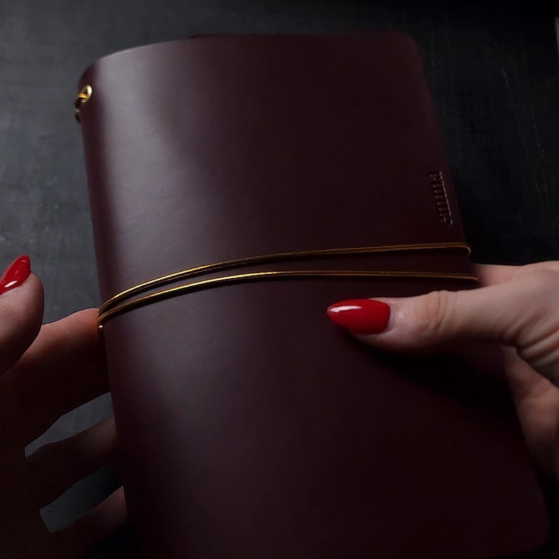 May include: A burgundy leather journal with a gold elastic closure. The journal is held by a person with red nail polish. The journal has a simple, elegant design, suitable for writing or sketching.