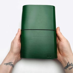 May include: A dark green leather journal held by two hands. The journal is secured with a black elastic band. The word "Iris" is embossed on the lower right corner. The background is white.