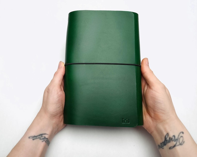 May include: A dark green leather journal held by two hands. The journal is secured with a black elastic band. The word "Iris" is embossed on the lower right corner. The background is white.