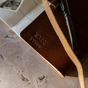 Puede incluir: Un diario de cuero marrón con cierre de correa y un bolígrafo. El diario tiene el texto "KMR 5 Years" grabado en relieve en la portada.