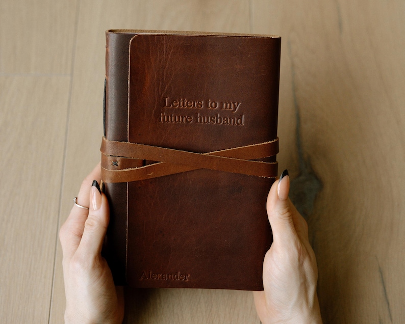 hands holding a 6x8.5" tan leather journal that is embossed with "letters to my future husband" in the upper middle. The name "alexander" in bottom corner of the flap. Tan is a reddish brown. The journal has a wrap around closure
