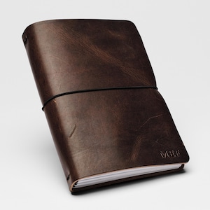 May include: A dark brown leather journal with a smooth, distressed texture and visible grain. The cover is secured by a thin black elastic band wrapped horizontally around the center. The letters MEE are embossed in the bottom right corner of the cover.