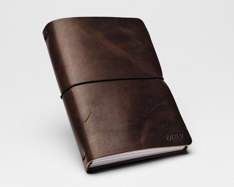 May include: A dark brown leather journal with a smooth, distressed texture and visible grain. The cover is secured by a thin black elastic band wrapped horizontally around the center. The letters MEE are embossed in the bottom right corner of the cover.