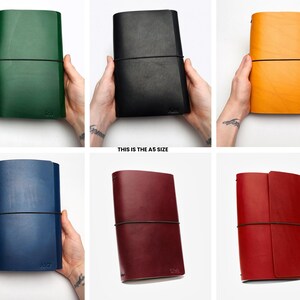 May include: A collection of A5 size leather journals in various colors: green, black, yellow, blue, burgundy, and red. Each journal is held open with a black elastic band. The text "THIS IS THE A5 SIZE" is visible.