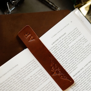Peut inclure: Un marque-page en cuir marron avec un motif d'oiseau et les initiales "AF" en relief sur le devant. Le marque-page est placé sur un livre avec le titre "A Mystery Revealed" visible.