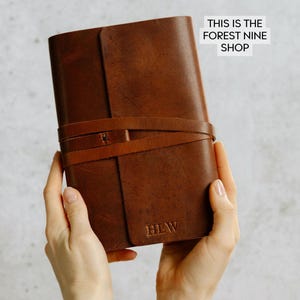 May include: A brown leather journal with a strap closure and the initials "HLW" embossed on the front. The journal is held in the hands of a person. "This is the Forest Nine Shop" is written in white text on a white background.