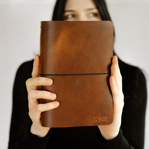 May include: A rectangular brown leather journal featuring a smooth, matte finish and a thin black elastic band wrapped horizontally around the center. The bottom right corner of the cover is embossed with the capital letters KNR in a serif font.