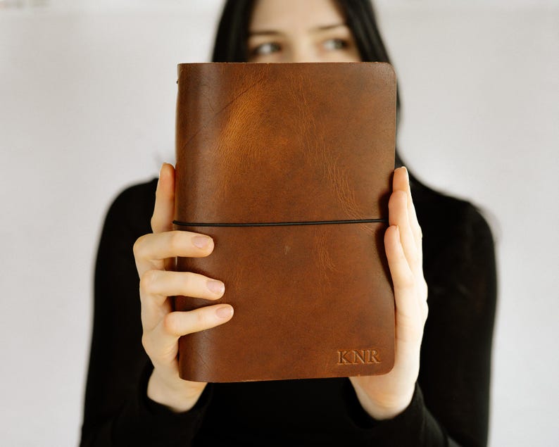 May include: A rectangular brown leather journal featuring a smooth, matte finish and a thin black elastic band wrapped horizontally around the center. The bottom right corner of the cover is embossed with the capital letters KNR in a serif font.