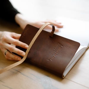 Pode incluir: Um di&aacute;rio de couro marrom com uma al&ccedil;a de couro bege e a inscri&ccedil;&atilde;o "Suas hist&oacute;rias para inspirar" na capa. O di&aacute;rio est&aacute; aberto em uma p&aacute;gina em branco.