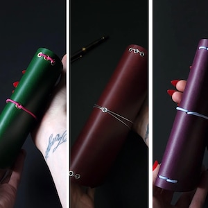May include: Three leather journal cases in green, burgundy, and purple. Each case is cylindrical and secured with a contrasting cord. The green case has pink cord, the burgundy has silver wire, and the purple has white cord.
