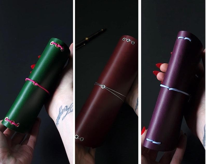 May include: Three leather journal cases in green, burgundy, and purple. Each case is cylindrical and secured with a contrasting cord. The green case has pink cord, the burgundy has silver wire, and the purple has white cord.