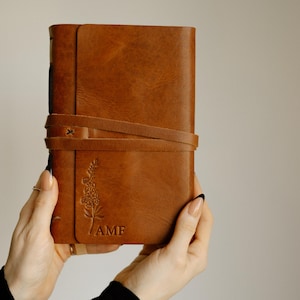 May include: A brown leather journal with a floral design and the initials "AMF" embossed on the front cover. The journal has a leather strap closure.