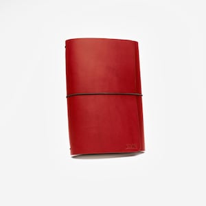 May include: A red leather journal with a dark brown elastic closure. The journal is rectangular and has rounded corners. The initials "LMR" are embossed on the bottom right corner. The journal is set against a white background.