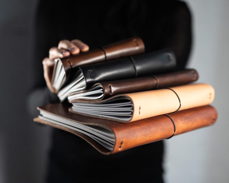 May include: A stack of five leather-bound journals in various shades of brown, black, and tan. Each notebook features a flexible leather cover, visible stitching along the spine, and a thin black elastic band wrapped around the center to secure the pages.