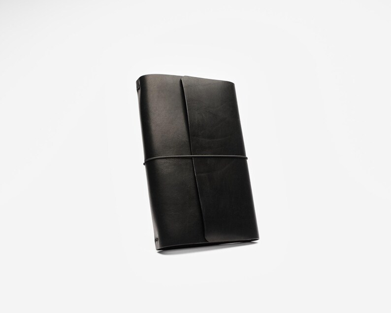 May include: A black leather journal with a wraparound elastic closure. The journal is rectangular and appears to be closed. The leather has a matte finish. The journal is set against a white background.