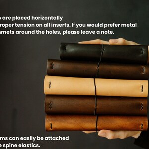 May include: A stack of leather journals in various shades of brown and tan, held by a person. The journals are bound with black elastic and feature horizontal holes. Text on the image mentions the option for metal grommets and charm attachment.