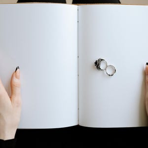 Pode incluir: Duas alian&ccedil;as de prata, uma com um design detalhado, s&atilde;o colocadas em um livro branco aberto. O livro &eacute; mantido aberto por duas m&atilde;os com esmalte preto nas unhas.