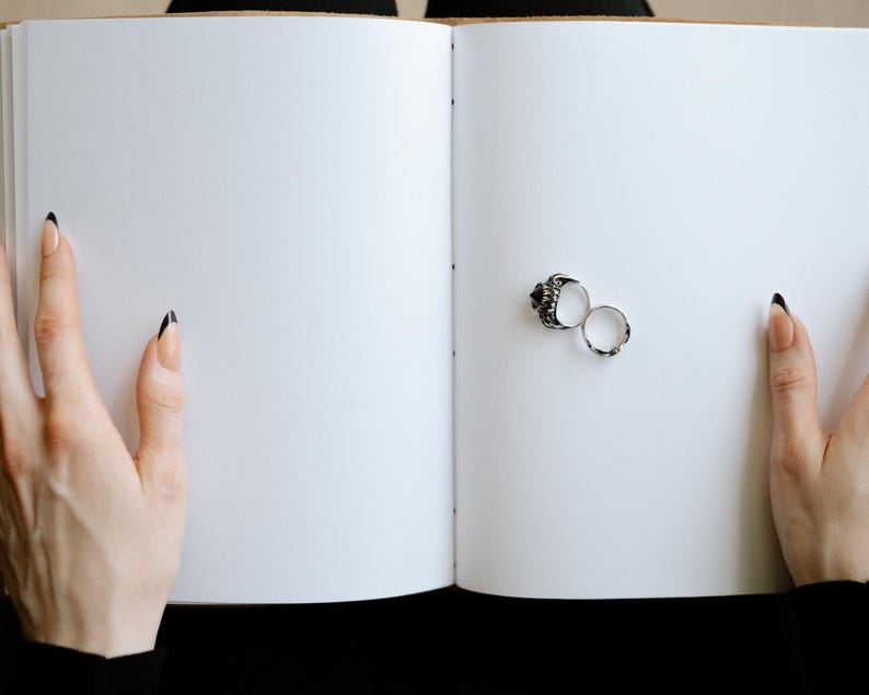 Pode incluir: Duas alian&ccedil;as de prata, uma com um design detalhado, s&atilde;o colocadas em um livro branco aberto. O livro &eacute; mantido aberto por duas m&atilde;os com esmalte preto nas unhas.