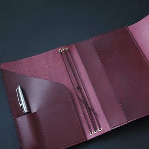 Op de afbeelding: Open bordeaux leren notitieboek met meerdere vakken en elastische sluitingen. Een zilveren pen zit in de voorzak. Het notitieboek is open en onthult de binnenpagina's en stikdetails. De achtergrond is donker.