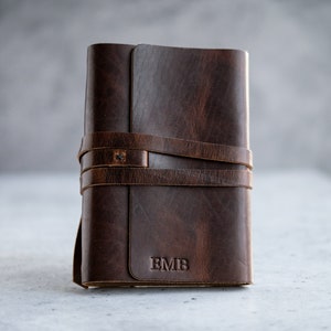 May include: A brown leather journal with a wrap-around strap closure. The journal features the initials "EMB" embossed on the front cover. The leather has a distressed appearance, and the strap is secured with a small metal rivet.