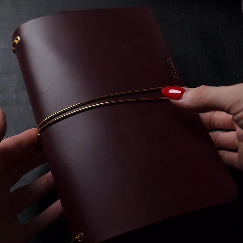 May include: A burgundy leather journal with a gold elastic closure. The journal is closed, with the word "timeless" embossed on the side. The journal is held by a person with red nail polish.