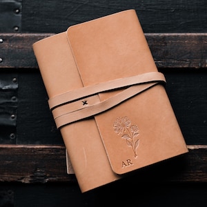 May include: A light brown leather journal with a strap closure. The cover features an embossed floral design and the initials "AR". The journal is closed with a wrap-around leather strap.