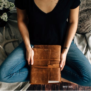 May include: A brown leather journal with the text "MYB" embossed on the front cover. The journal is closed and has a strap closure. The journal is being held by a person wearing blue jeans and a black shirt.