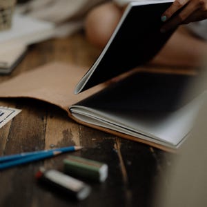 May include: A tan leather notebook cover holding a black and white journal, accompanied by two blue pencils and two rectangular erasers. A blue and white printed paper document with architectural lines is visible on the left side of the frame.
