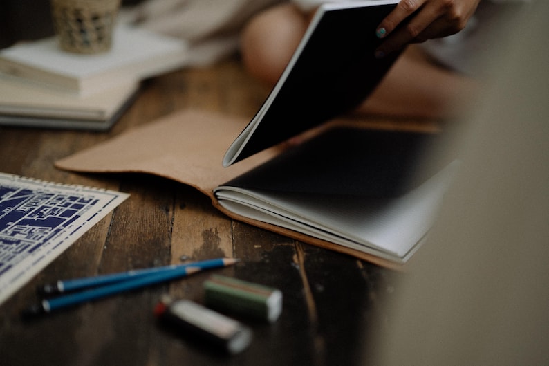 May include: A tan leather notebook cover holding a black and white journal, accompanied by two blue pencils and two rectangular erasers. A blue and white printed paper document with architectural lines is visible on the left side of the frame.