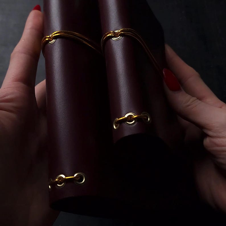 May include: Two burgundy leather journals with gold-colored elastic closures. The journals are held together with a gold elastic band. The image is a close-up shot, highlighting the texture and color of the leather.