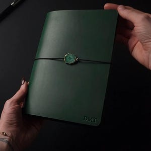 Peut inclure: Un carnet en cuir vert foncé avec une finition mate lisse et les lettres DMT embossées dans le coin inférieur droit. Un fin cordon élastique noir entoure horizontalement la couverture, maintenu par une breloque en agate verte.