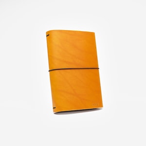 May include: A golden-yellow leather journal with a black elastic band closure. The journal has a minimalist design with the initials "AMP" embossed on the bottom right corner. The cover is smooth and the edges are rounded.