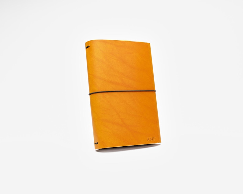 May include: A golden-yellow leather journal with a black elastic band closure. The journal has a minimalist design with the initials "AMP" embossed on the bottom right corner. The cover is smooth and the edges are rounded.