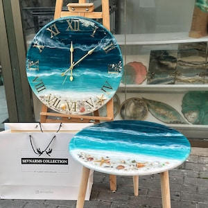 May include: A round wall clock and a matching round side table, both featuring a resin ocean wave design with turquoise and deep blue hues, white foam accents, and embedded seashells. The clock has gold Roman numerals and gold hands.