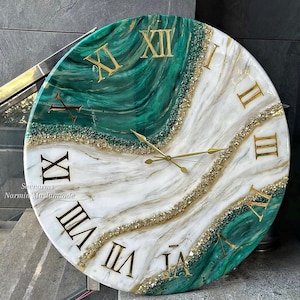 Puede incluir: Un reloj de pared redondo con un diseño jaspeado en verde esmeralda y blanco con números romanos dorados. Una banda diagonal de purpurina dorada texturizada y acentos de piedra triturada divide los colores. Tres finas manecillas doradas están centradas.