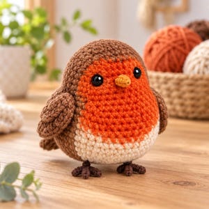 May include: A small, round, crocheted bird figurine featuring a brown back and wings, a bright orange chest, and a cream-colored belly. The bird has two black plastic eyes, a small yellow beak, and dark brown crocheted feet standing upright.