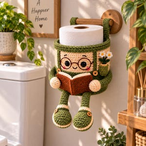 May include: A crochet toilet paper holder shaped like a character with glasses, wearing green overalls, and holding a brown book. The holder features a small pocket with white flowers, dangling legs with green shoes, and is mounted on a wooden rod.