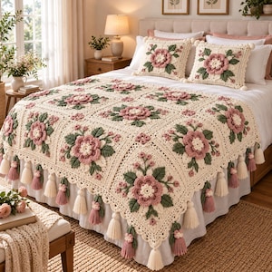 May include: A cream-colored crochet bedspread and two matching square throw pillows featuring a raised floral pattern with dusty pink petals, white centers, and sage green leaves. The bedspread edges are finished with decorative hanging tassels.