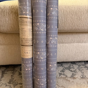 May include: Three rolls of grey wallpaper featuring a repeating pattern of small white flowers and thin horizontal lines. The leftmost roll includes a tan label with the text Wallcovering, 18756, and RUN-2, indicating a bolt size of 33 inches by 20.5 inches.