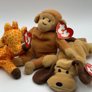 May include: Three plush toys including an orange and yellow giraffe with a spotted pattern, a light brown monkey, and a tan dog with dark brown ears. Each stuffed animal features a red heart-shaped Ty tag attached to its ear or body.
