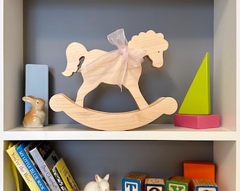 May include: A wooden rocking horse with a pink ribbon bow sits on a shelf. Other items on the shelf include a ceramic bunny, colorful blocks, and small books. The word "TOYS" is spelled out with wooden blocks.