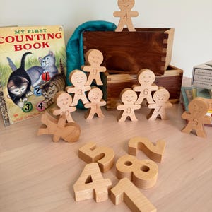 May include: A collection of light-colored wooden figures shaped like gingerbread people, each engraved with a unique number from one to ten. The set includes several standing figures, one lying down, and separate wooden number blocks arranged in front.