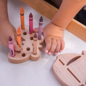 May include: A wooden toy with a bear shape and holes for crayons. A child is holding a pink crayon and inserting it into one of the holes. There are other crayons in the holes, including orange, pink, purple, and brown.