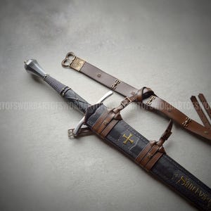 Puede incluir: Una espada de acero con empuñadura estriada y pomo metálico, junto a una vaina negra con un emblema de cruz dorada y correas de cuero marrón. La vaina incluye detalles metálicos decorativos y un largo cinturón de cuero marrón.