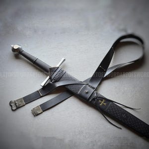 May include: A longsword with a silver pommel and crossguard, housed in a black leather scabbard featuring an embossed geometric pattern, a gold cross emblem, and a black leather belt strap with metallic hardware. The text ARTOFSWORD is visible.