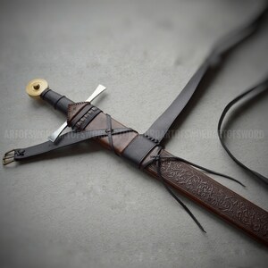 May include: A sword with a black leather-wrapped hilt, a circular brass pommel, and a straight silver crossguard. The brown leather scabbard features intricate embossed floral patterns, black leather straps, and a metal buckle attachment.