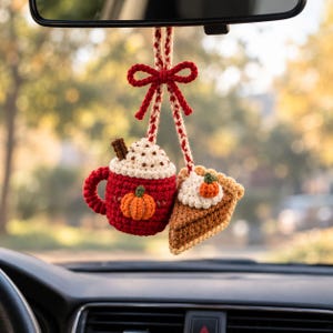 Può includere: Un accessorio per auto all'uncinetto composto da una tazza rossa con una zucca e panna montata, abbinata a una fetta di torta di zucca con panna, entrambi appesi a un cordino intrecciato rosso e bianco legato con un fiocco decorativo in cima.