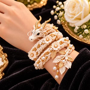 May include: A cream-colored crochet dragon bracelet featuring a coiled body, gold-toned horns, and intricate floral accents. The design is embellished with small white pearls and metallic gold beads, finished with a cluster of gold-toned leaf charms.