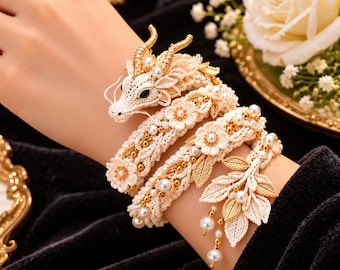 May include: A cream-colored crochet dragon bracelet featuring a coiled body, gold-toned horns, and intricate floral accents. The design is embellished with small white pearls and metallic gold beads, finished with a cluster of gold-toned leaf charms.